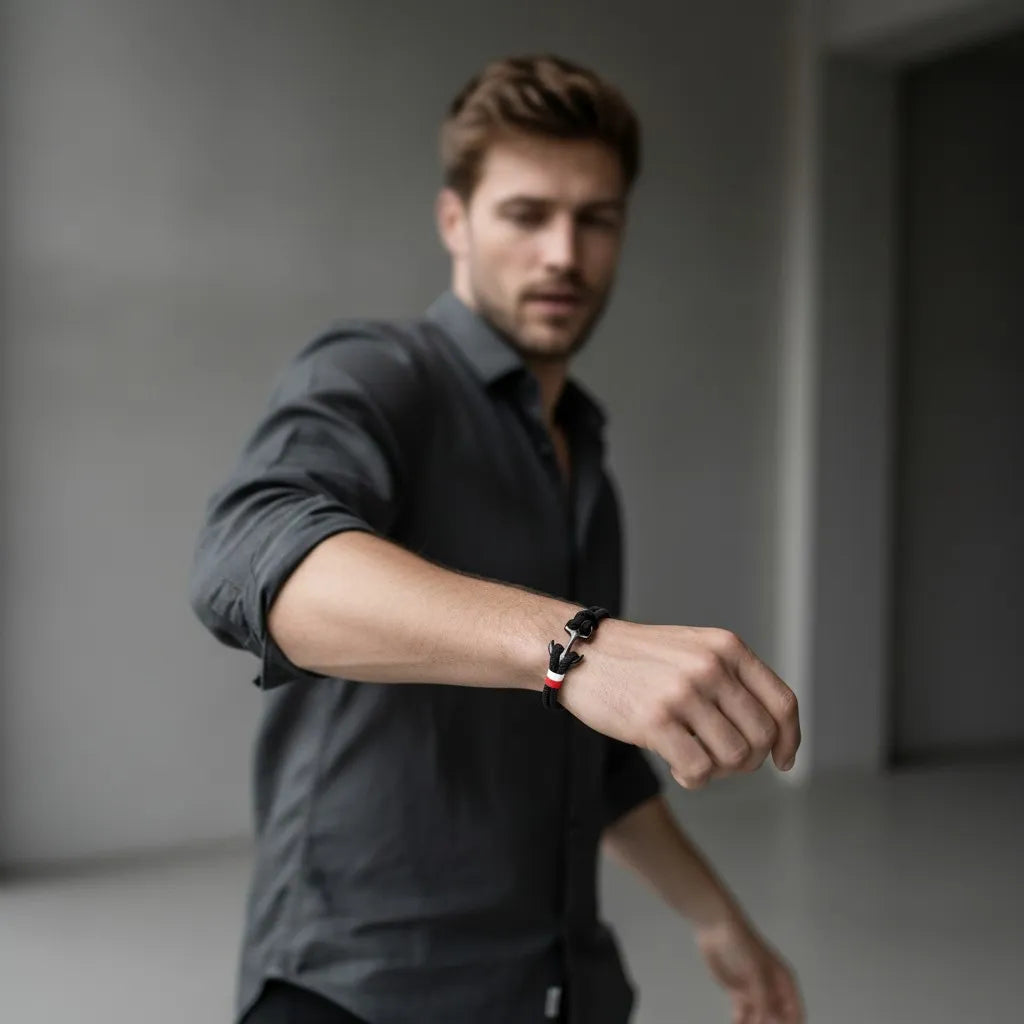 Homem em pé com braço estendido usando pulseira de corda preta com detalhe metálico escuro em ambiente interno minimalista.