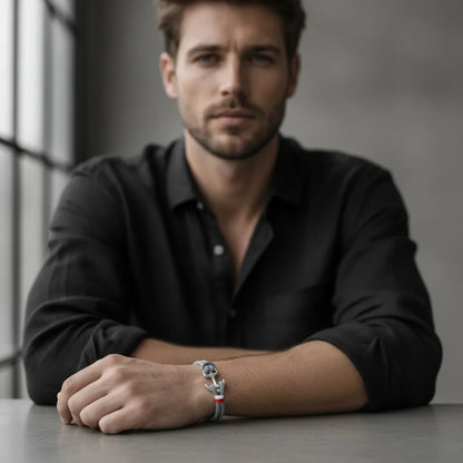 Modelo masculino sentado à mesa usando pulseira de corda cinza com detalhe metálico prateado no pulso.