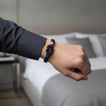 Pulseira masculina de corda azul-marinho com nó e detalhes tricolores no pulso, em um ambiente interno minimalista.