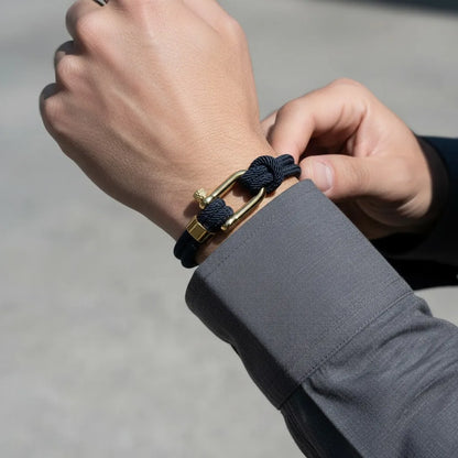 Homem ajustando pulseira masculina de corda azul com nós aparentes e fecho metálico dourado no pulso.