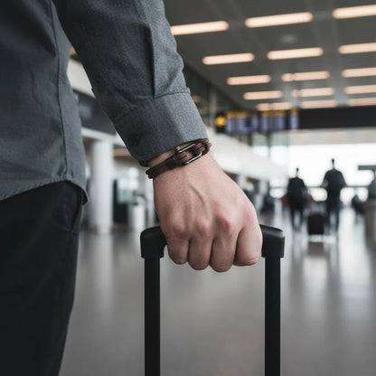 Homem usando pulseira masculina de corda marrom com fecho preto enquanto segura a alça de uma mala em ambiente de aeroporto.