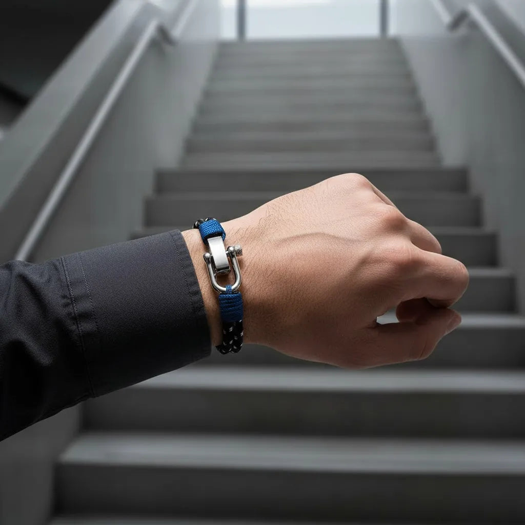 Homem usando pulseira masculina de corda preta com detalhes azuis e fecho metálico diante de uma escada em ambiente moderno.