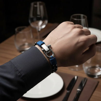 Close da pulseira masculina de corda preta com detalhes azuis e fecho metálico no pulso, sobre mesa posta em ambiente elegante.