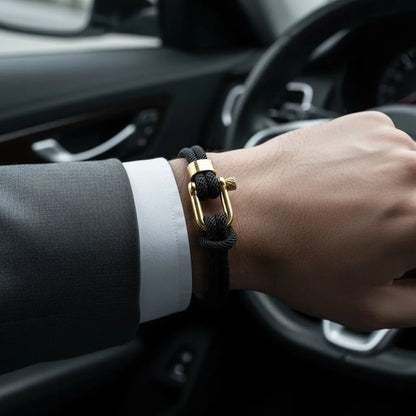 Pulseira masculina de corda preta com fecho metálico dourado usada no pulso dentro de um carro.