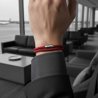 Close da pulseira masculina de corda vinho com volta dupla e fecho metálico prateado em um ambiente moderno de sala de espera.