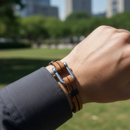 Pulseira masculina de couro marrom com detalhe metálico prateado sendo usada no pulso em ambiente ao ar livre.
