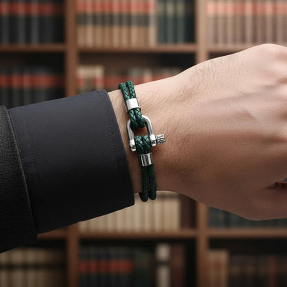 Pulseira masculina de couro verde trançado com fecho metálico em destaque diante de uma estante de livros.