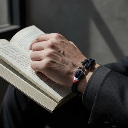 Pulseira de corda azul com âncora em aço no pulso enquanto a pessoa segura um livro aberto.
