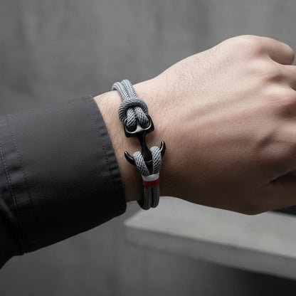 Close no pulso masculino usando pulseira de corda cinza com âncora preta e detalhe em vermelho e branco, em fundo cinza minimalista.
