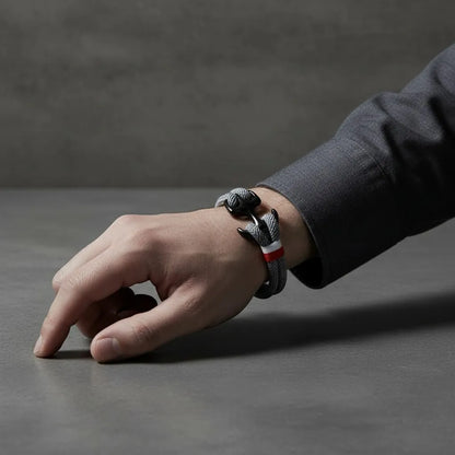 Pulso masculino apoiado sobre mesa cinza usando pulseira de corda cinza com âncora preta e detalhe em vermelho e branco.