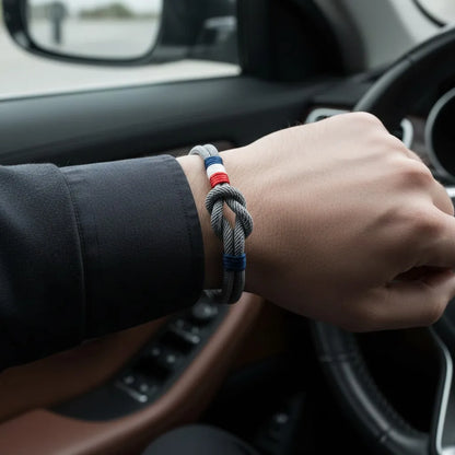 Pulseira de corda cinza com nó central e detalhes tricolores em vermelho, branco e azul, usada no pulso dentro de um carro, com volante ao fundo.