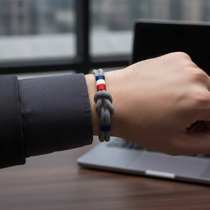 Pulseira de corda cinza com nó central e detalhes tricolores em vermelho, branco e azul, usada no pulso em frente a uma mesa de trabalho com notebook ao fundo.