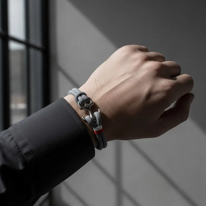 Close no pulso masculino usando pulseira de corda cinza com detalhe metálico prateado e faixa vermelha, em fundo minimalista com luz natural.