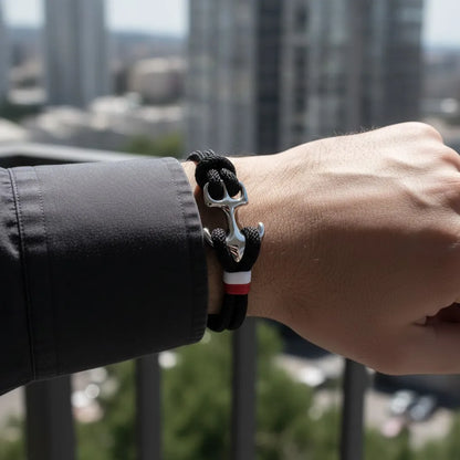 Close do pulso com pulseira de corda preta com âncora em aço, com prédios ao fundo.