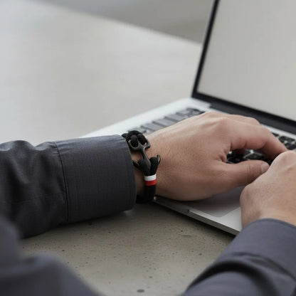 Pulso masculino com pulseira de corda preta e detalhe metálico escuro em destaque, enquanto digita no notebook.