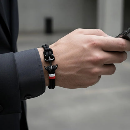 Close no pulso masculino usando pulseira de corda preta com detalhe metálico escuro e faixa branca e vermelha, enquanto segura um celular.