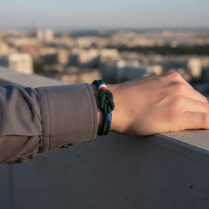 Pulseira de corda em tom verde escuro com nó central e detalhes tricolores em vermelho, branco e azul, usada no pulso apoiado sobre uma mureta com vista desfocada da cidade ao fundo.