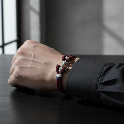 Close no pulso masculino usando pulseira de corda vinho com detalhe metálico prateado e faixa branca e azul, apoiado sobre mesa preta.