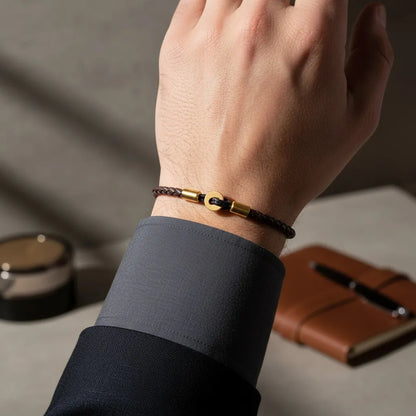 Pulseira de couro marrom com detalhes dourados usada no pulso, iluminada por luz natural, com caderno e caneta ao fundo.