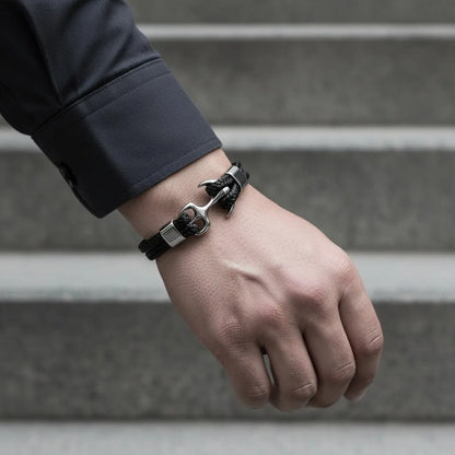 Pulseira de couro preta com âncora em metal prateado no pulso masculino, fotografada diante de uma escada cinza.