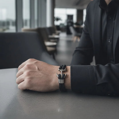 Pulseira de couro preta com âncora em metal prateado no pulso masculino apoiado sobre uma mesa em ambiente corporativo.