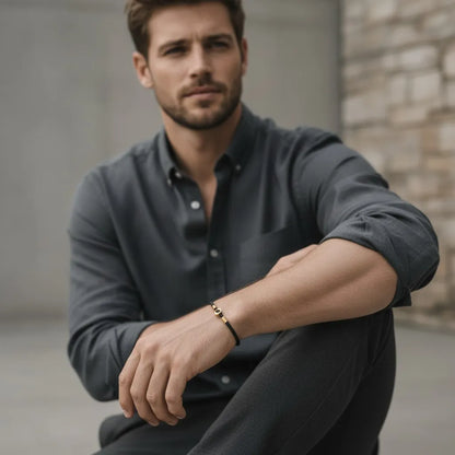 Pessoa sentada usando pulseira de couro preta com detalhes dourados, combinada com camisa escura em um visual casual elegante.