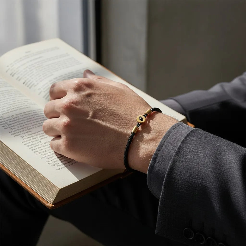 Pulseira de couro preta com detalhes dourados usada no pulso enquanto a pessoa segura um livro aberto.
