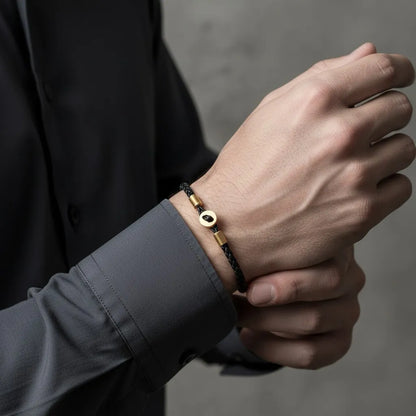 Pulseira de couro preta com detalhes dourados sendo usada no pulso, com a outra mão apoiando o braço em fundo neutro.