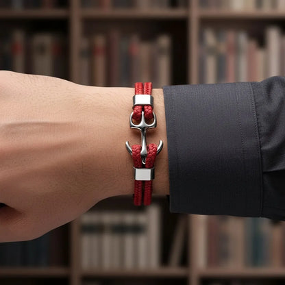 Close frontal da pulseira de couro vermelha com âncora em metal prateado no pulso, com estante desfocada ao fundo.