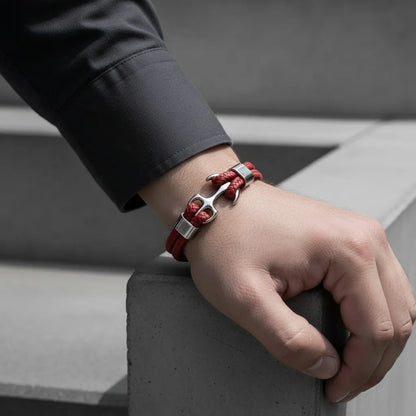 Close no pulso masculino usando pulseira de couro vermelha com âncora em metal prateado, apoiado sobre estrutura de concreto.
