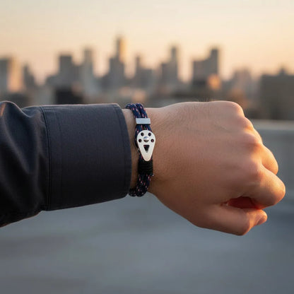 Pulseira masculina de corda azul com fecho metálico prateado em close no pulso, com fundo urbano ao entardecer.