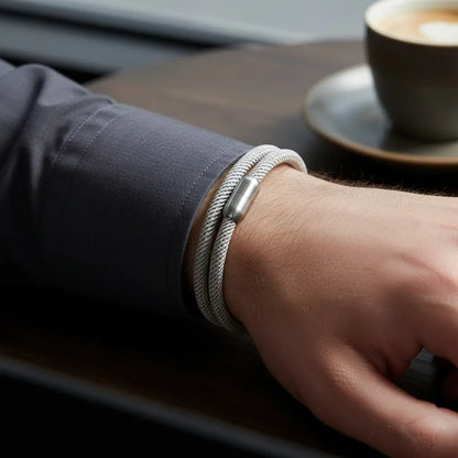 Pulseira masculina de corda cinza com fecho metálico prateado em close, usada no pulso sobre uma mesa ao lado de uma xícara de café.