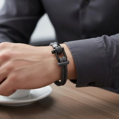 Close de pulseira masculina de corda cinza com fecho preto no pulso ao lado de uma xícara de café.