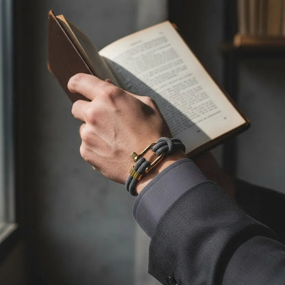 Homem lendo um livro enquanto usa pulseira masculina de corda cinza com fecho dourado em ambiente sofisticado.