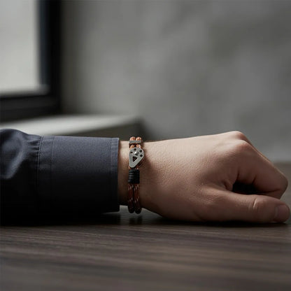 Pulseira masculina de corda marrom com detalhes pretos e peça metálica prateada, usada no pulso apoiado sobre uma mesa.