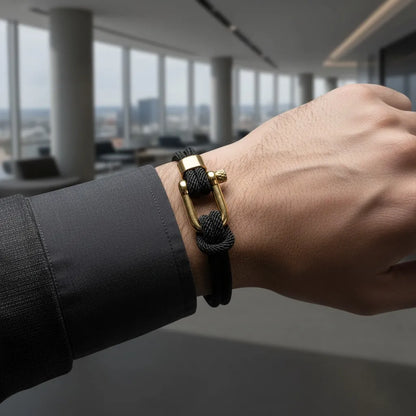 Close da pulseira masculina de corda preta com design sofisticado e fecho dourado em um ambiente corporativo.