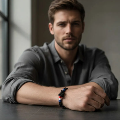 Homem sentado usando pulseira masculina de corda preta com nó e detalhes coloridos em um visual casual e elegante.