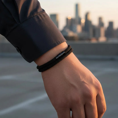Pulseira masculina de corda preta no pulso com paisagem urbana ao fundo.