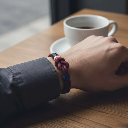 Close da pulseira masculina de corda vinho com nó central e detalhe azul no pulso, apoiado sobre uma mesa ao lado de uma xícara de café.