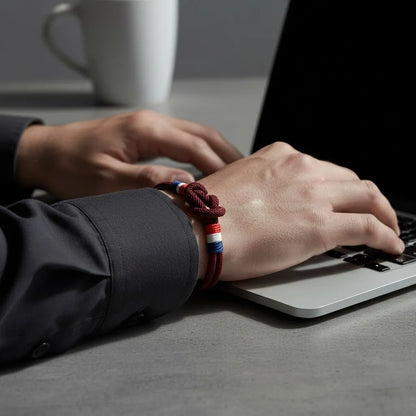 Pulseira masculina de corda vinho com nó central e detalhes em azul, branco e vermelho, usada no pulso enquanto a pessoa digita em um notebook.