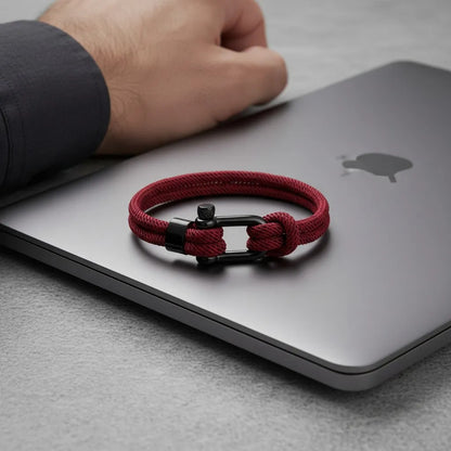 Pulseira masculina de corda vinho com fecho preto posicionada sobre um notebook fechado.