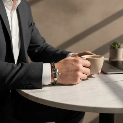 Homem de terno segurando uma xícara de café e usando pulseira masculina de couro cinza trançado com fecho metálico prateado.