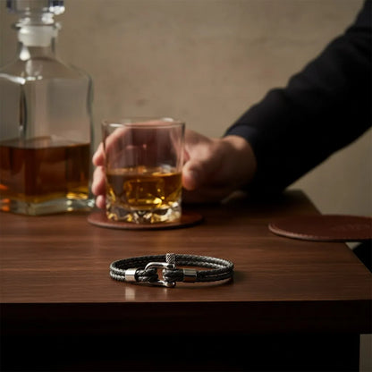 Pulseira masculina de couro cinza trançado com fecho metálico prateado sobre uma mesa de madeira ao lado de um copo.