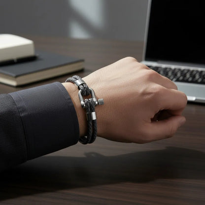 Detalhe de pulseira masculina de couro cinza trançado com fecho metálico prateado no pulso ao lado de um notebook.