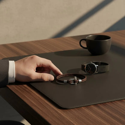 Pulseira masculina de couro marrom trançado com fecho metálico ao lado de relógio sobre mesa.