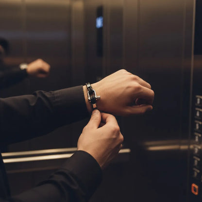Homem ajustando pulseira masculina de couro preto trançado com fecho prateado dentro de um elevador.
